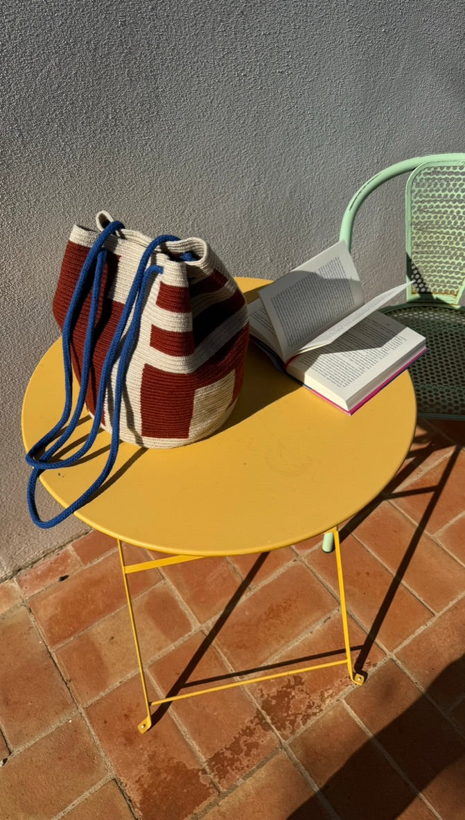 A red and beige colured knitted cotton bag with blue straps on a yellow table. An open book is on the table as well. 