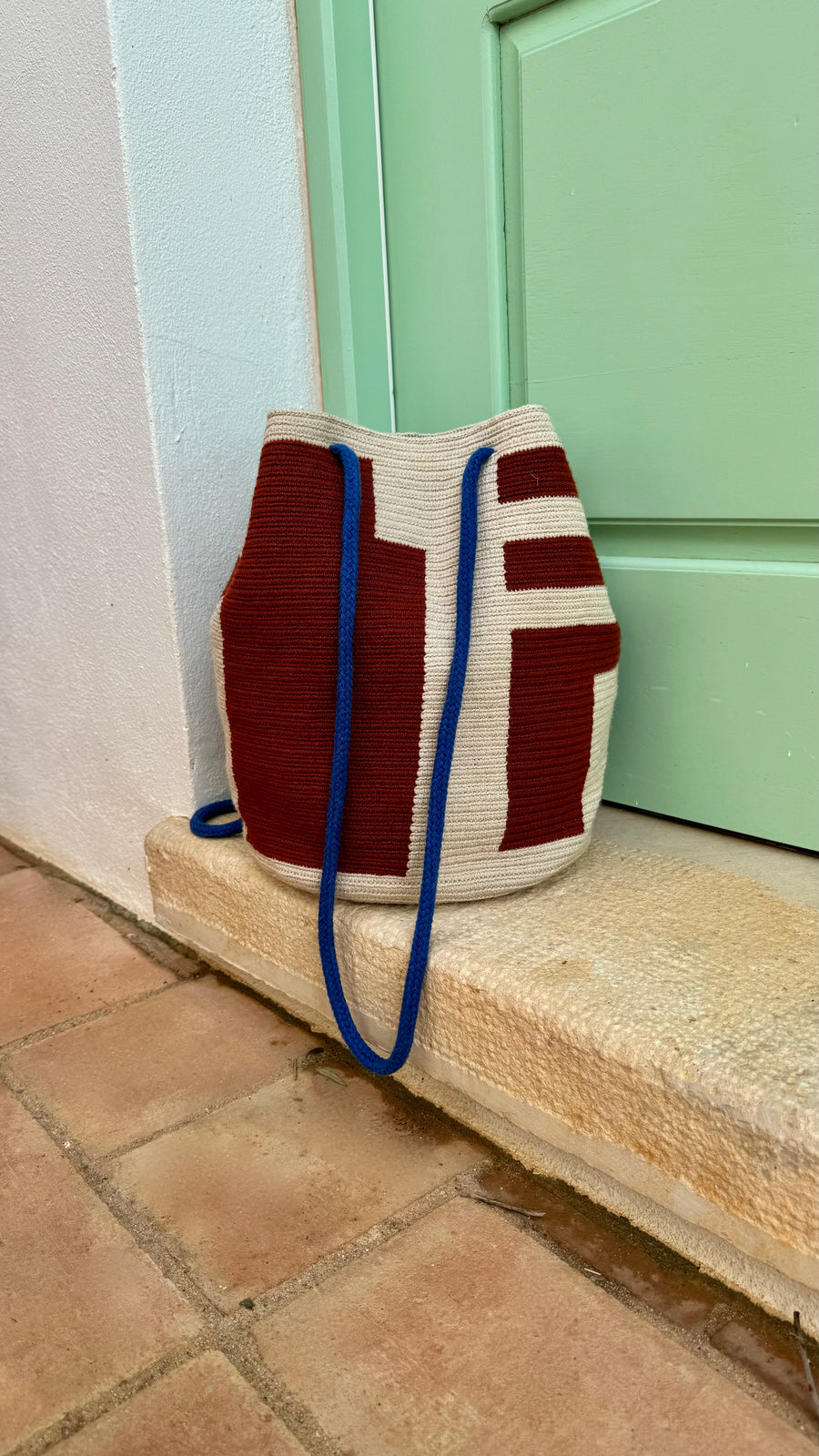 A red and beige coloured cotton bag with blue straps in front of a green coloured door